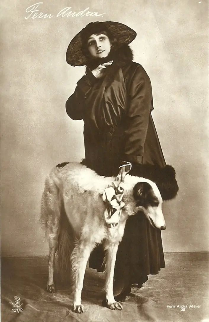 Postal fotográfica sepia: una actriz con sombrero de ala ancha y abrigo de piel, junto a un galgo ruso.
